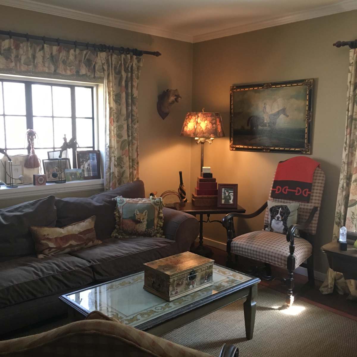 A cozy living room with a brown couch, a glass coffee table, and a patterned armchair beside a window adorned with floral curtains.