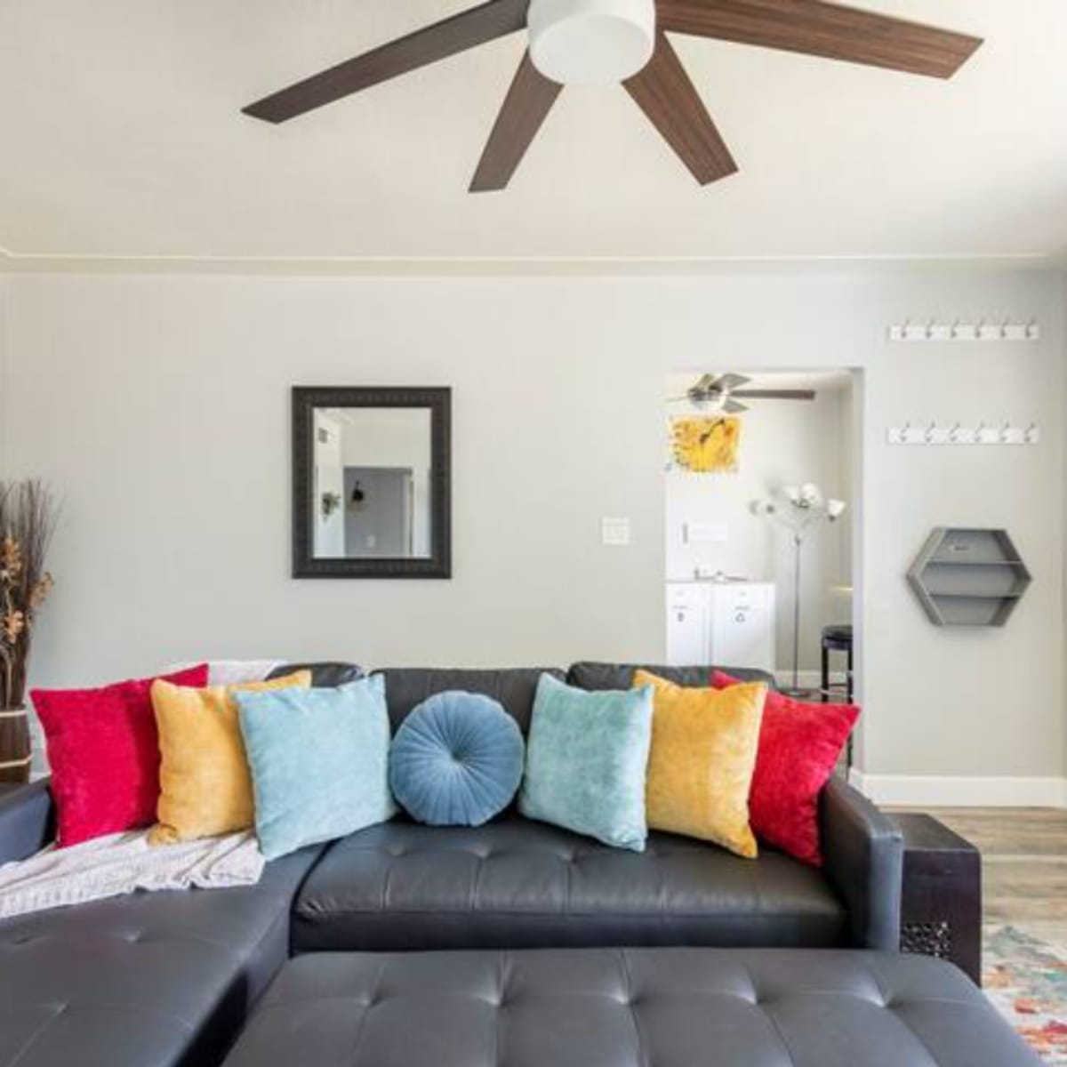 A black sectional sofa with colorful cushions is positioned in a well-lit living room featuring a ceiling fan and a doorway leading to a laundry area.