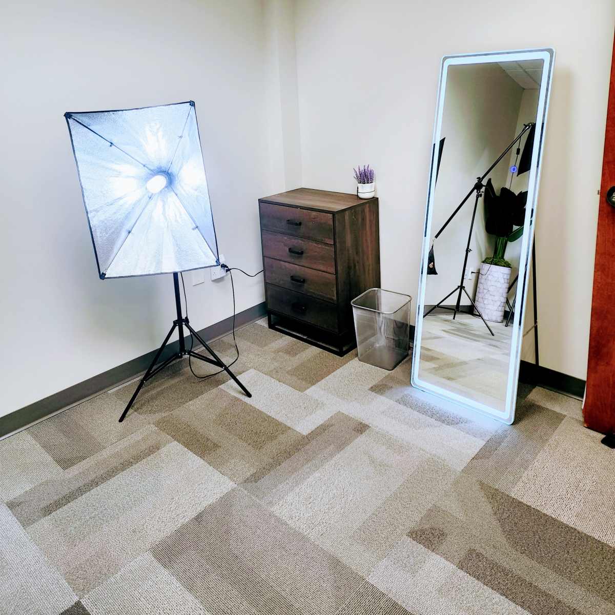 A corner of a room featuring a softbox light, a tall mirror, a small dresser with drawers, and a wastebasket.