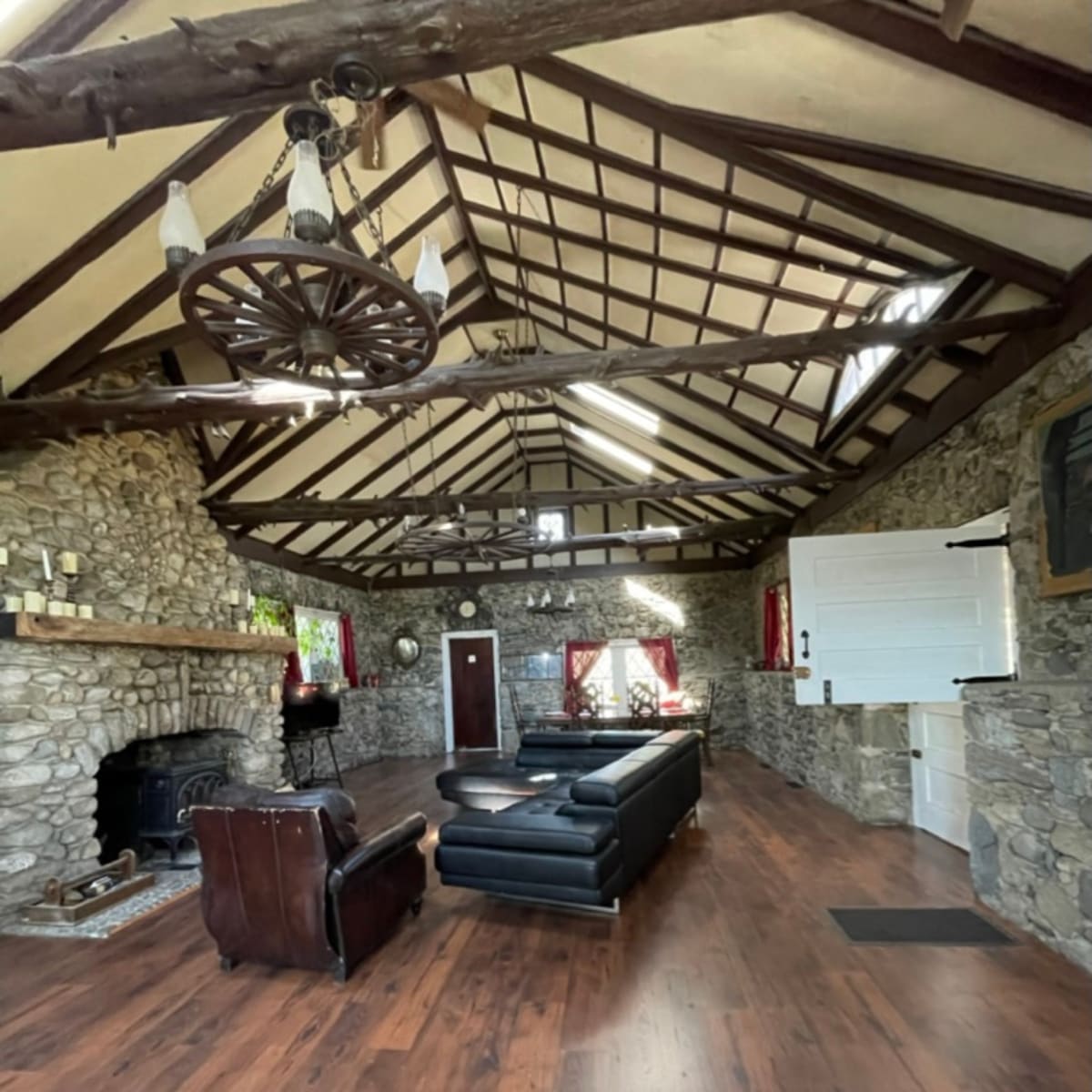 A spacious living room featuring stone walls, a high wooden ceiling with beams, and a black leather sofa centered on a wooden floor.