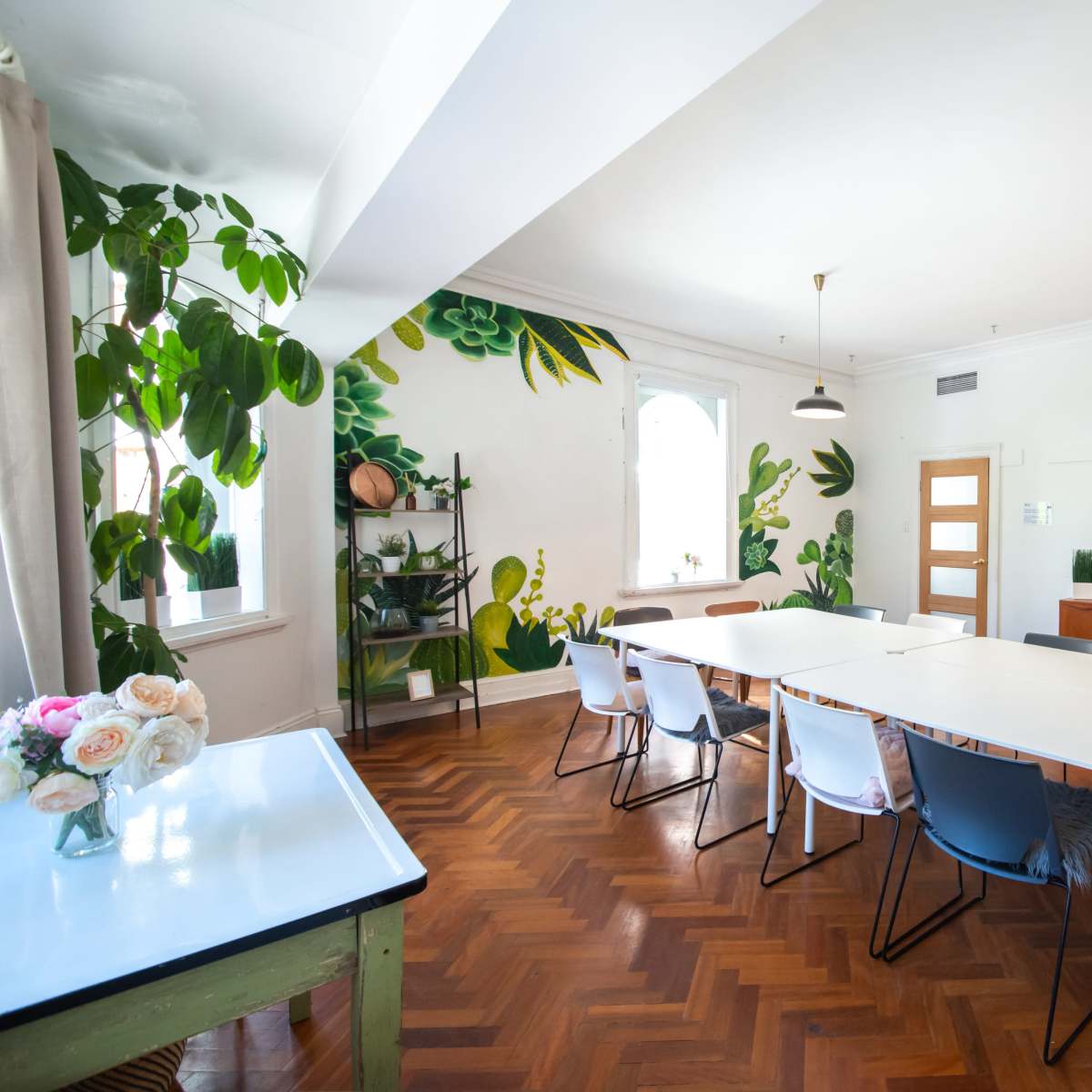 A well-lit meeting room with a long white table surrounded by chairs, a television on a stand, and a wall featuring a green leafy mural.