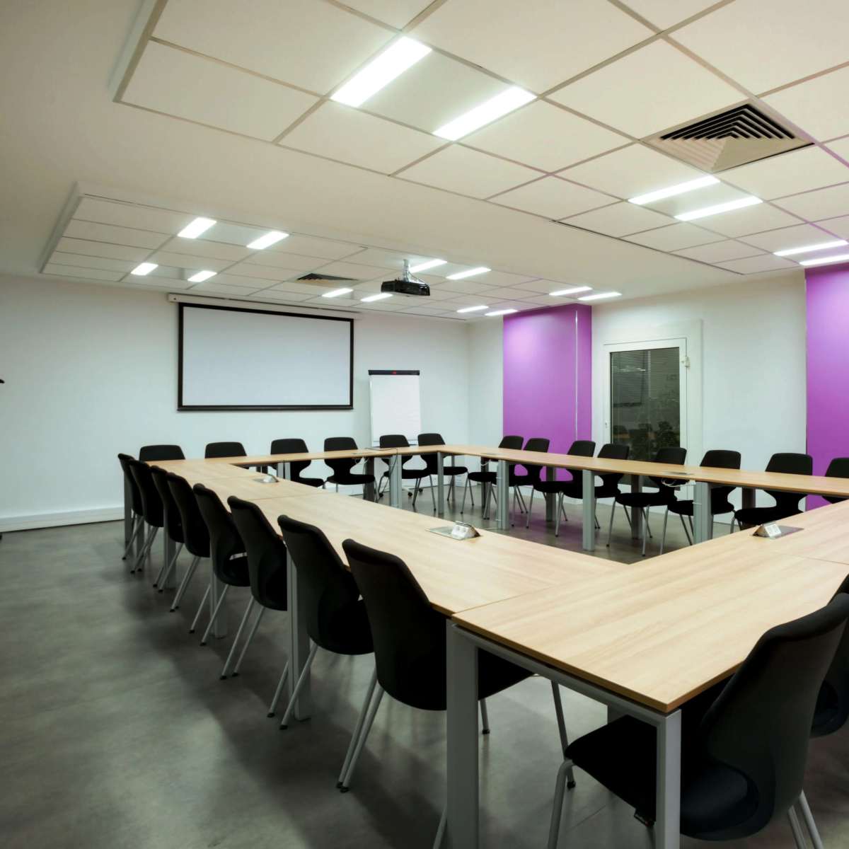 A conference room features a long, triangular table surrounded by black chairs, with a projector screen at the front and purple accent walls.