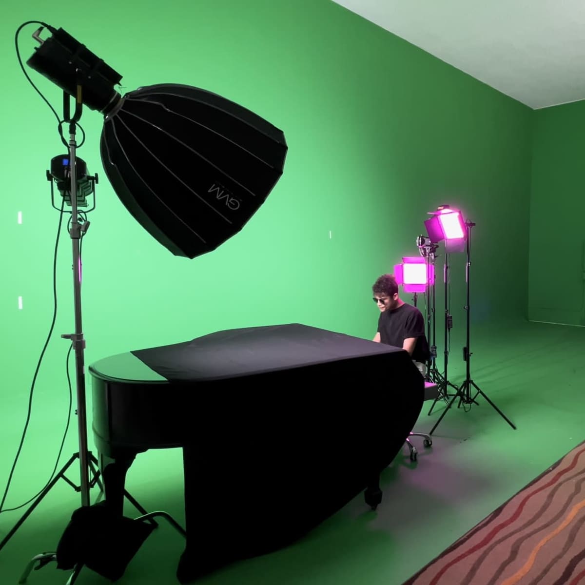 A person sits at a grand piano in a studio set up with green screens, softbox lighting, and additional colored lights.
