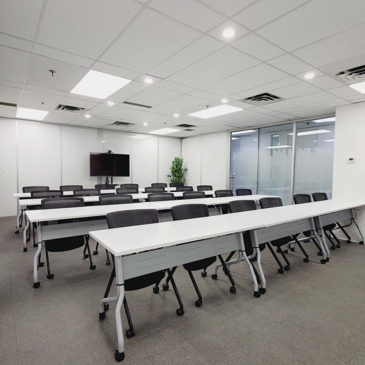 A modern conference room with rows of tables and chairs arranged neatly, featuring a large screen at one end and large windows allowing natural light.