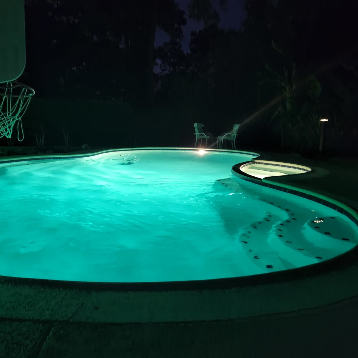 A swimming pool illuminated by green lights at night, with a basketball hoop and two chairs nearby.