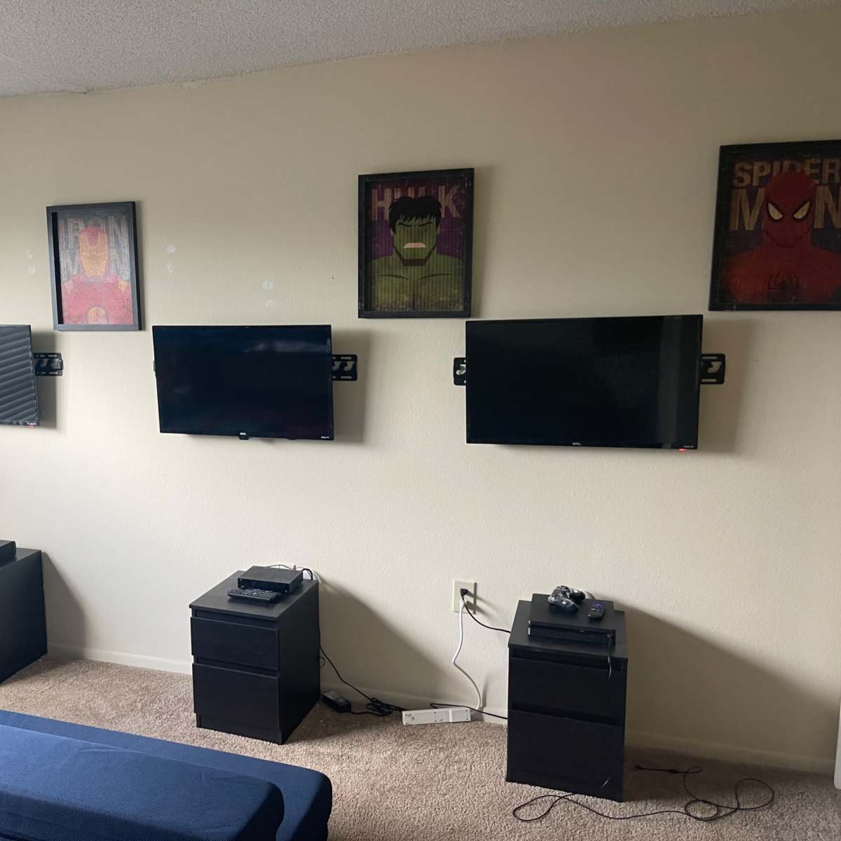 A living room wall with four mounted televisions flanked by framed superhero posters, and two black nightstands below each TV.