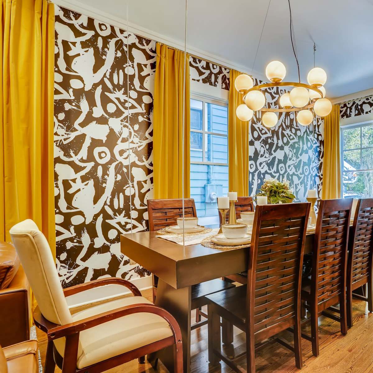 A dining room features a wooden table surrounded by chairs, with brown and white patterned wallpaper and yellow curtains framing the windows.