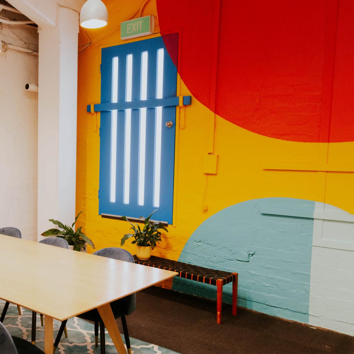 A bright meeting room features a large wooden table surrounded by six blue chairs, a wall-mounted television, and a colorful mural with a blue door and vibrant design.