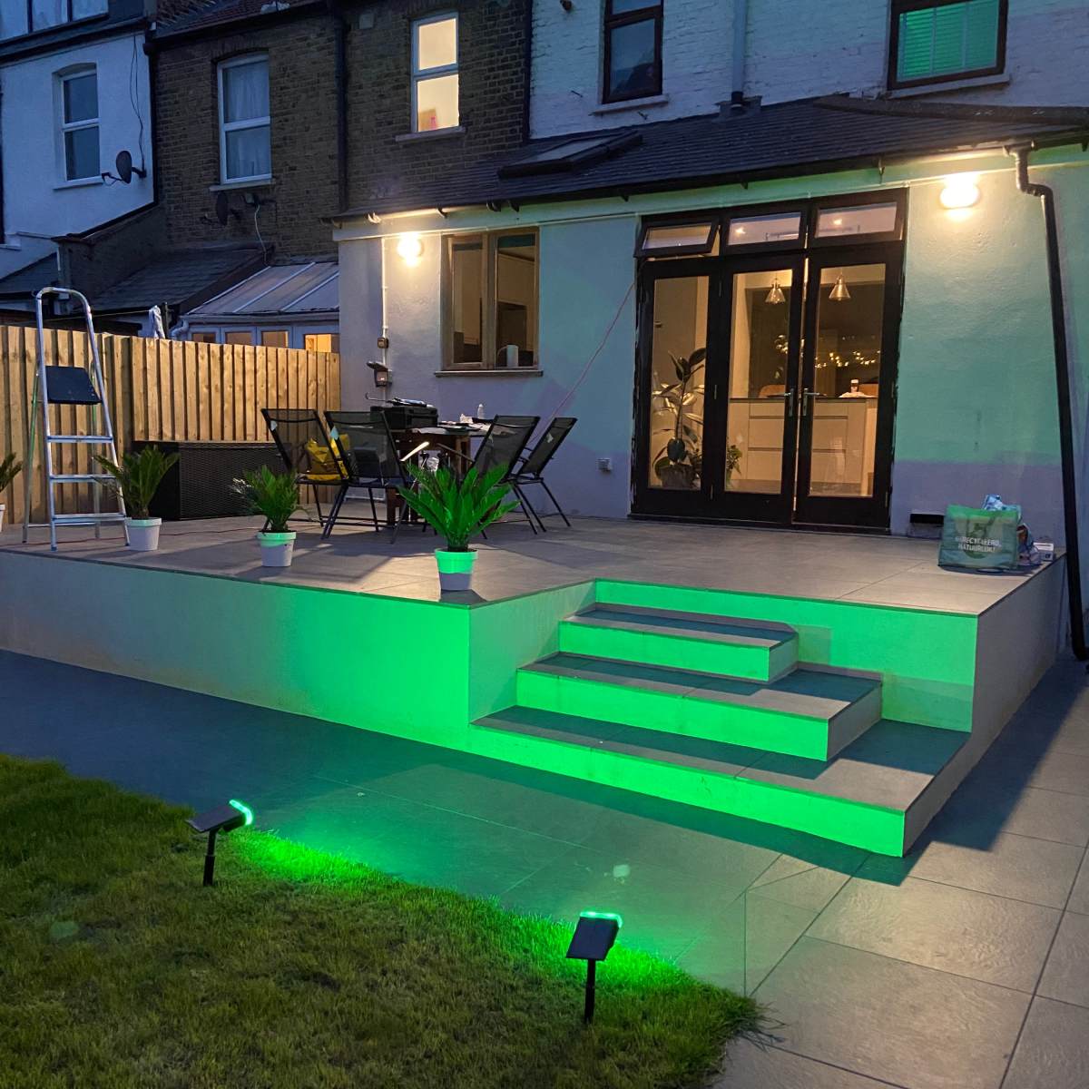 A backyard patio features green lighting, steps leading to the entrance, and seating arrangements near the house.