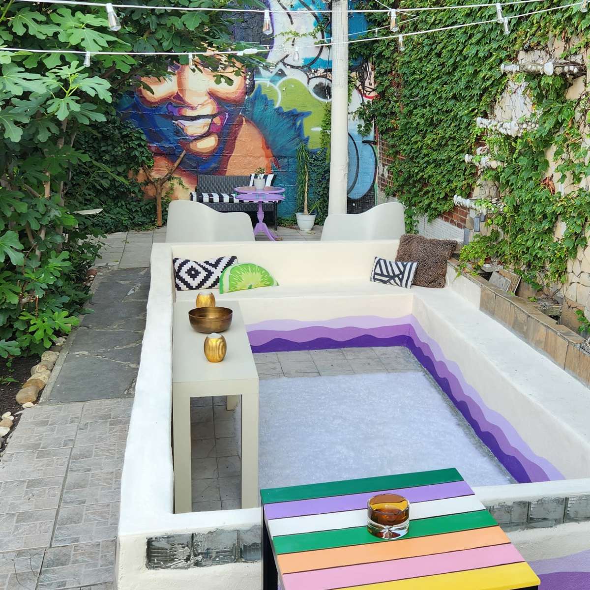 A colorful outdoor seating area with a mural, surrounded by greenery and featuring a unique, multicolored bench.