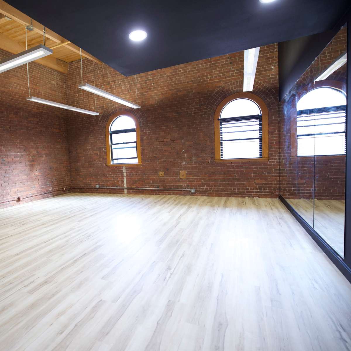 An empty room with exposed brick walls, wooden beams, and large arched windows, featuring a light-colored wooden floor and a mirrored wall.
