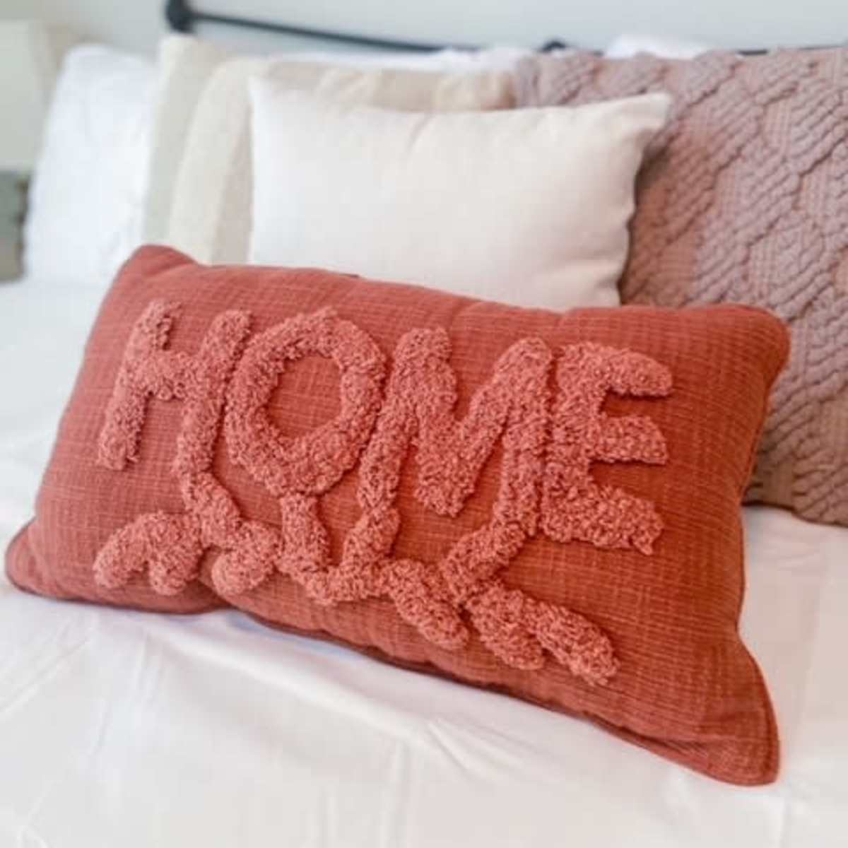 A large, textured pillow with the word "HOME" prominently displayed sits on a neatly made bed adorned with various other decorative pillows.