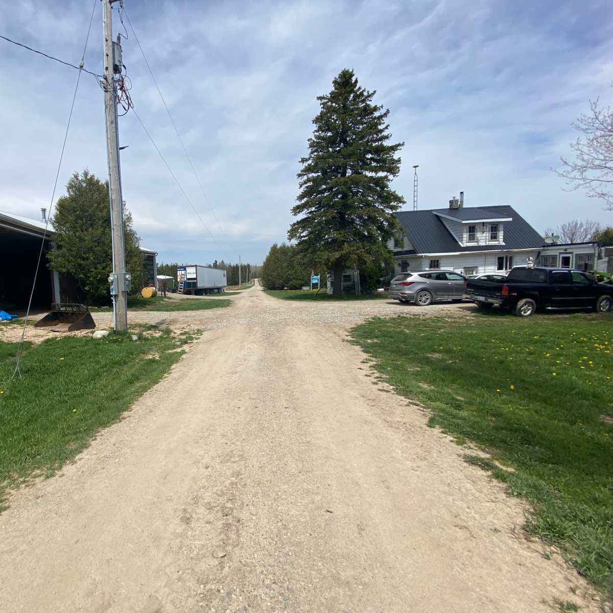 A dirt road leads through a rural area with two buildings on either side, flanked by trees and parked vehicles.