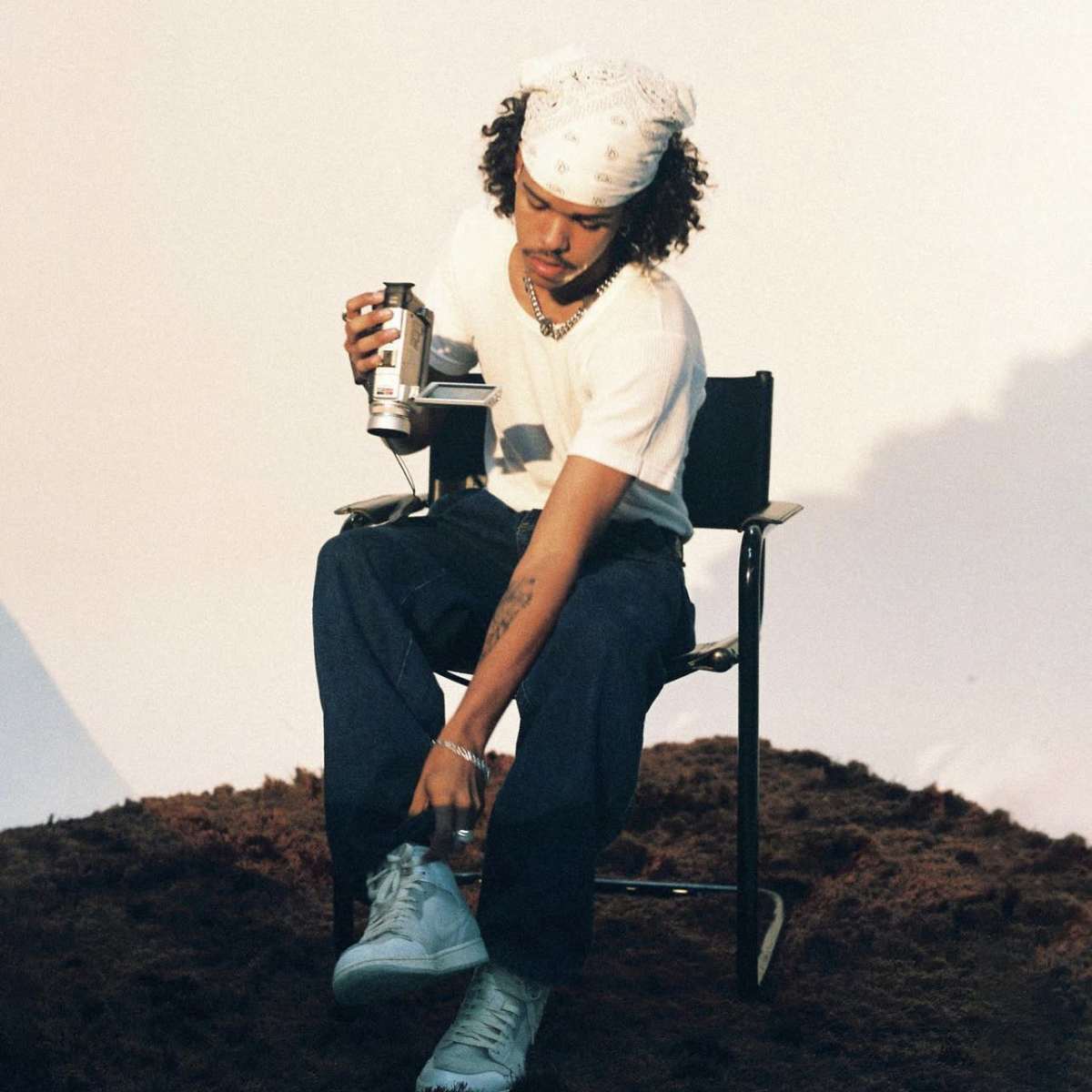 A person sitting on a chair adjusts their sneaker while holding a can and wearing a bandana.