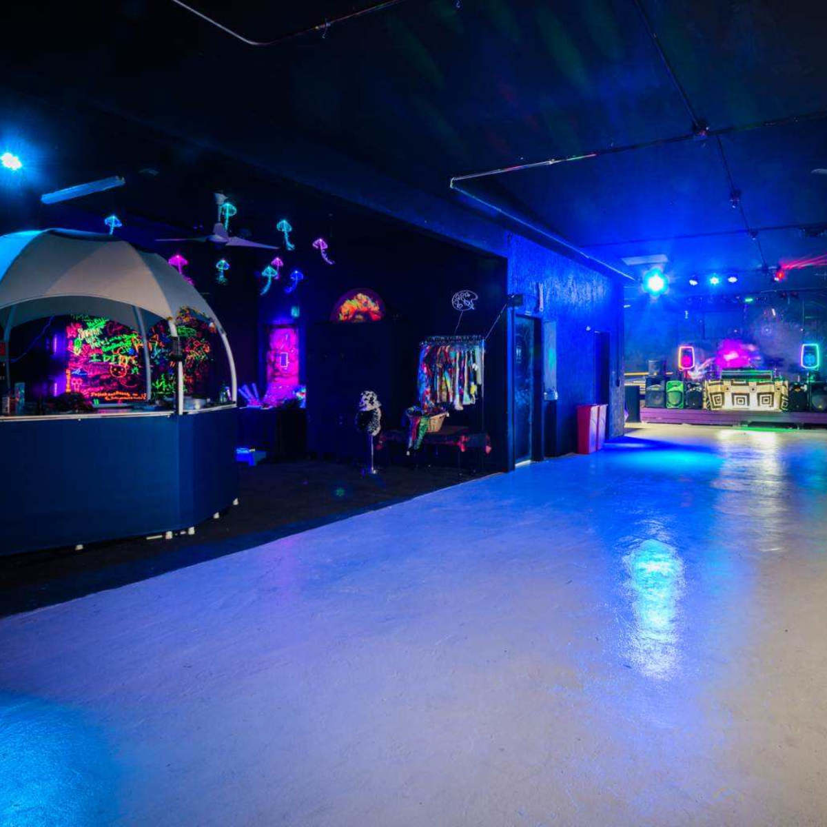 A spacious indoor venue featuring a brightly lit area with colorful neon decorations, a concession stand, and a DJ booth at the far end.