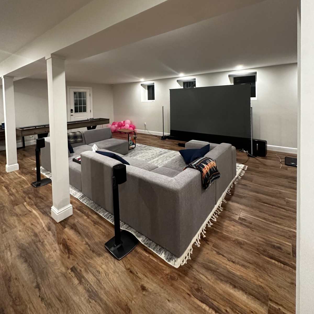 A spacious basement living area features a large gray sectional sofa facing a wall-mounted television, with wooden flooring and lit windows.