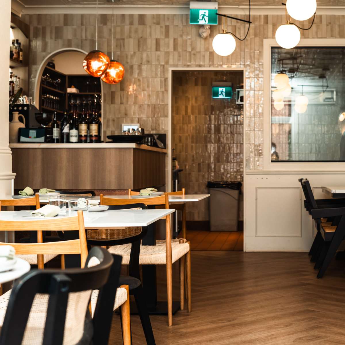 A modern restaurant interior with wooden tables, assorted chairs, and a bar area in the background.