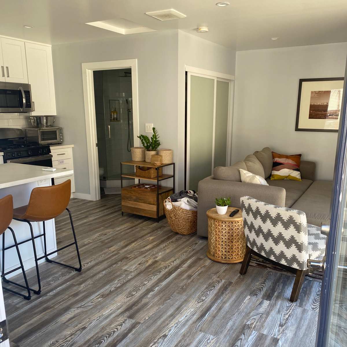 A modern living space with a gray sofa, wood flooring, and a kitchen area featuring two bar stools.