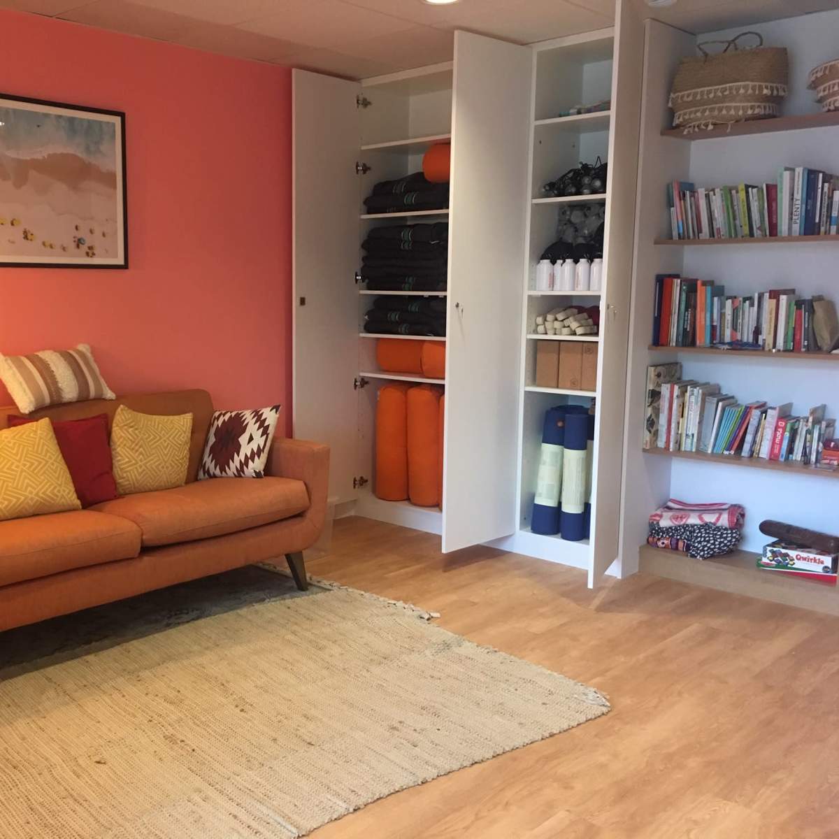 A bright living space features an orange sofa adorned with colorful pillows, a small side table, and a bookshelf filled with various books, alongside a storage cabinet.