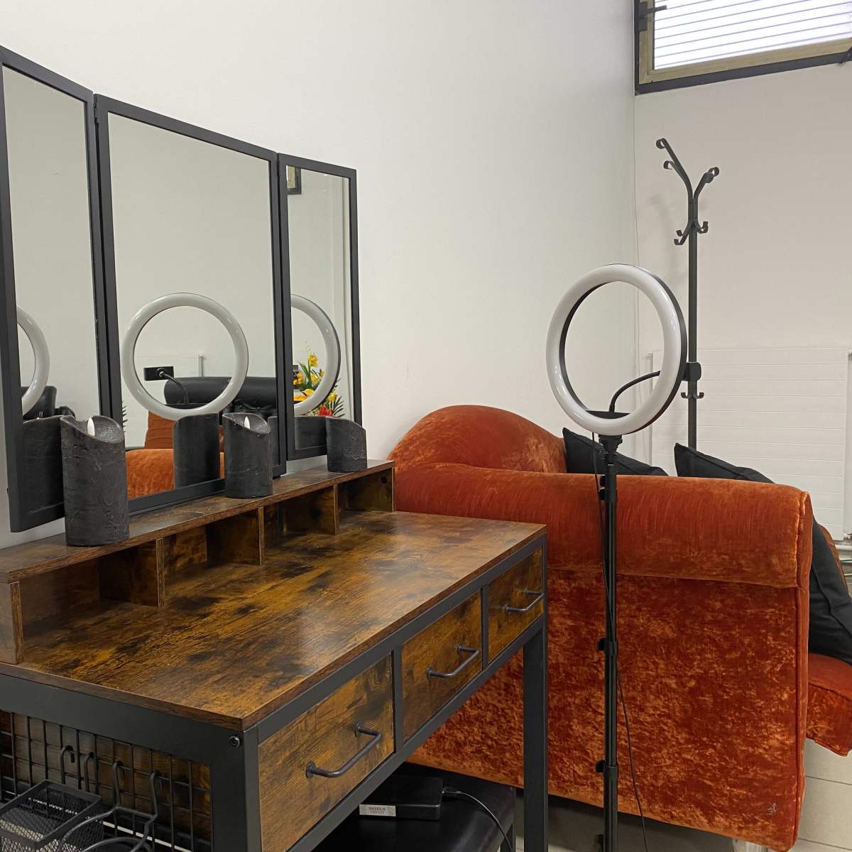 A wooden vanity with a three-panel mirror and decorative items is positioned next to an orange sofa in a minimalist room.