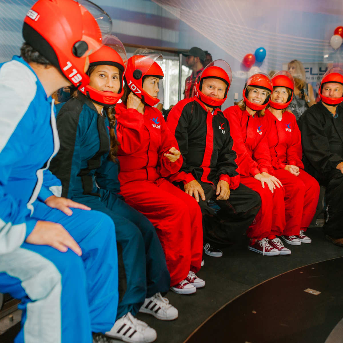 A group of people in colorful jumpsuits and helmets sits together, preparing for an indoor skydiving experience.