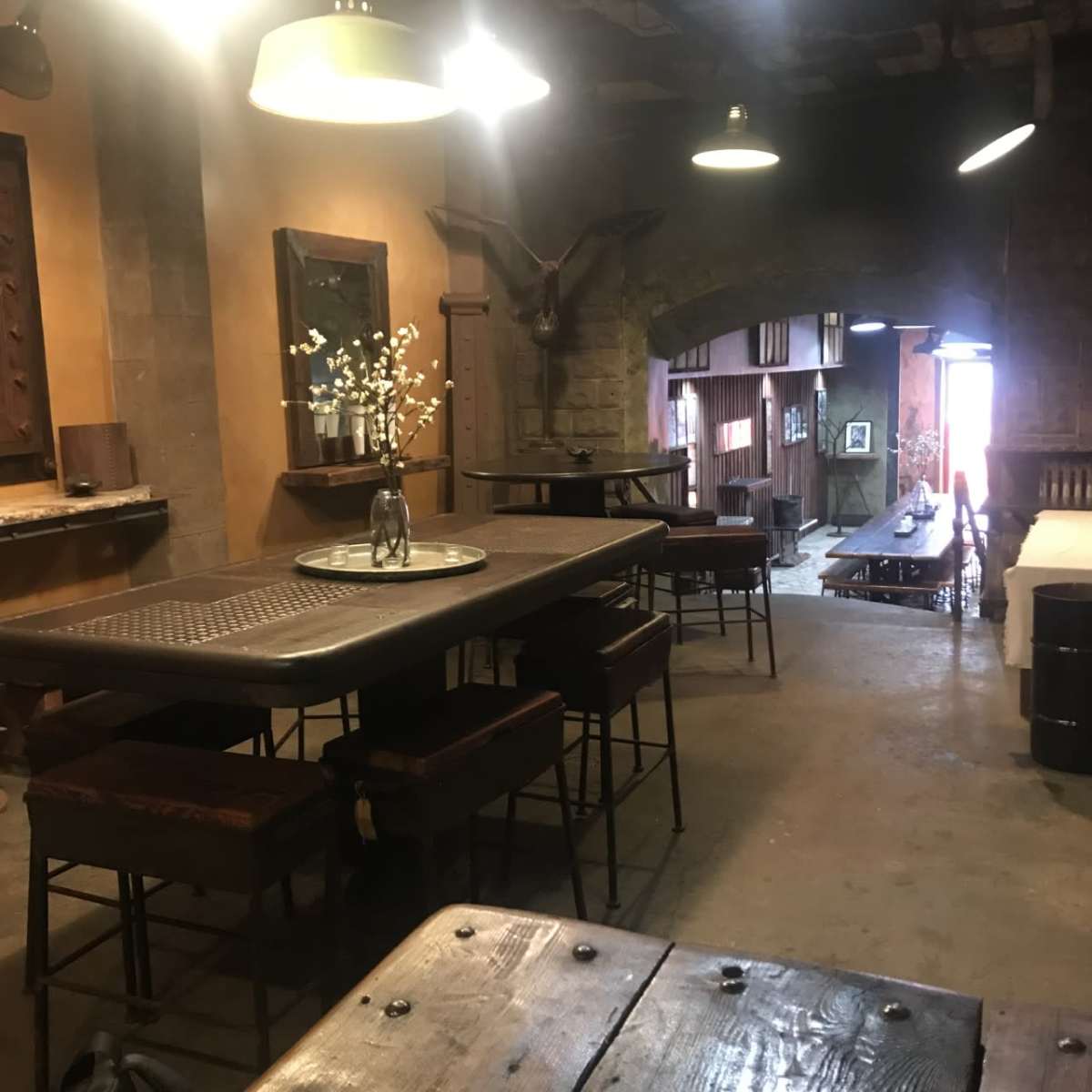A rustic interior dining area with wooden tables and chairs, featuring stone walls and low-hanging light fixtures.