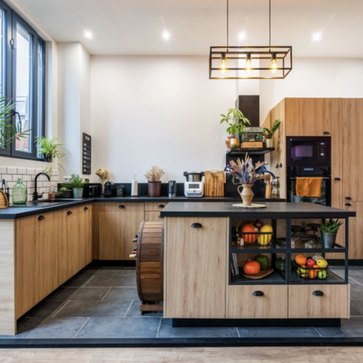 A modern kitchen featuring wooden cabinetry, a large island with black countertops, and various plants and fruits displayed throughout the space.
