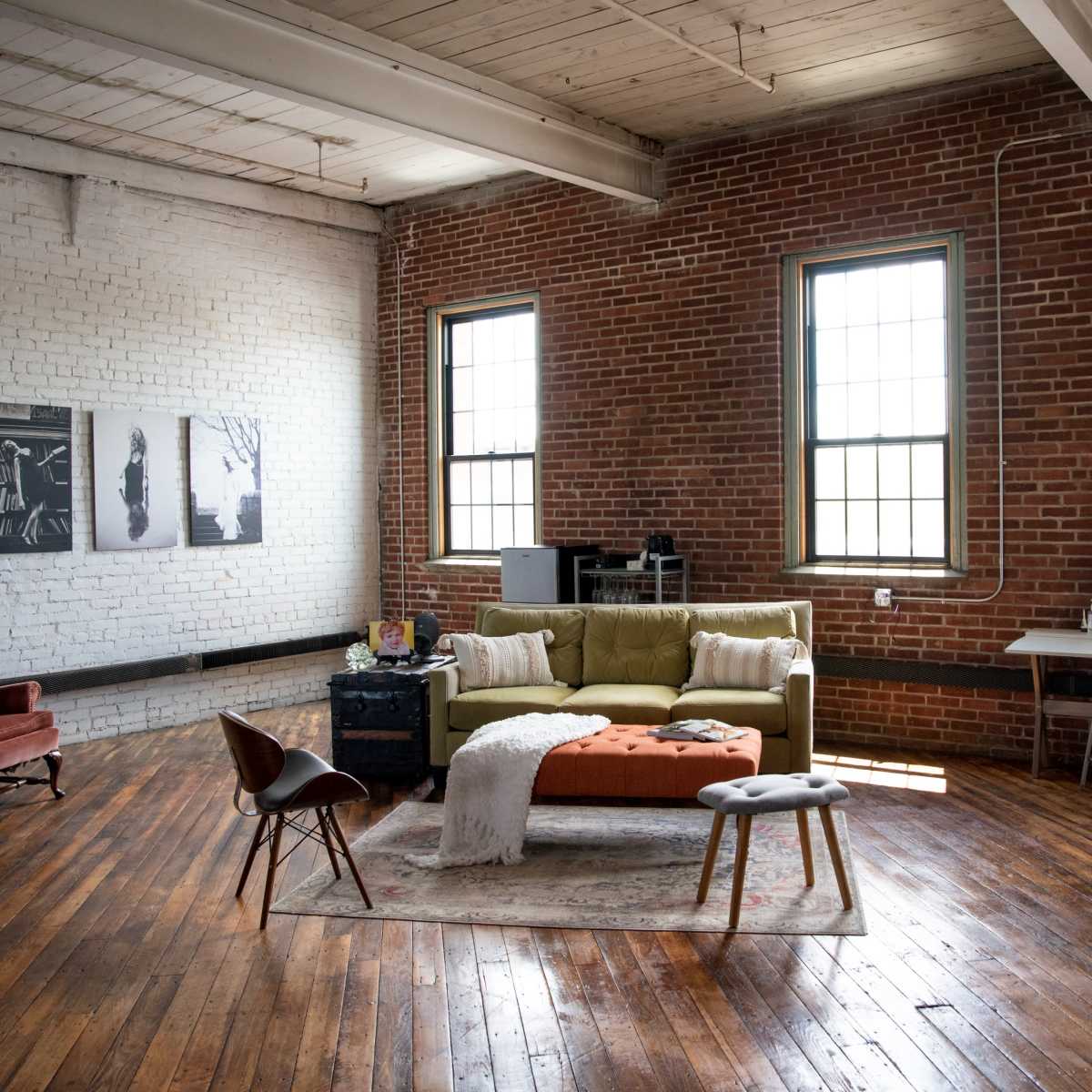 A spacious room with exposed brick walls, wooden floors, a green sofa, a rust-colored ottoman, and a small desk near two large windows.