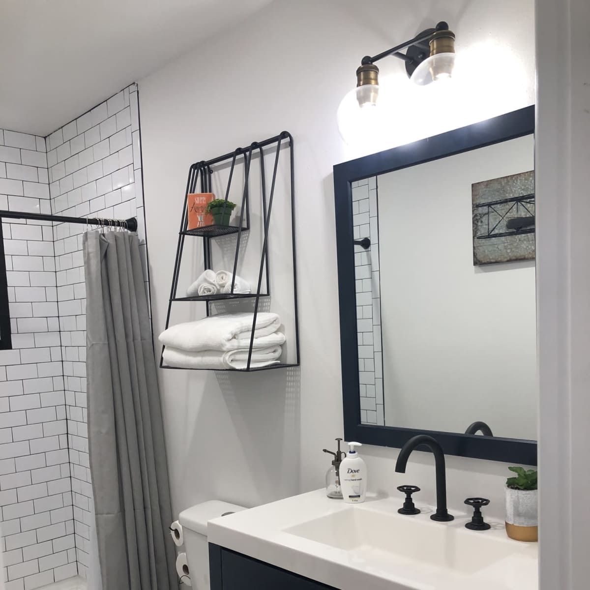 A modern bathroom featuring a white-tiled shower area, a black vanity with a sink and faucet, a mirror, and a metal shelf holding towels and plants.