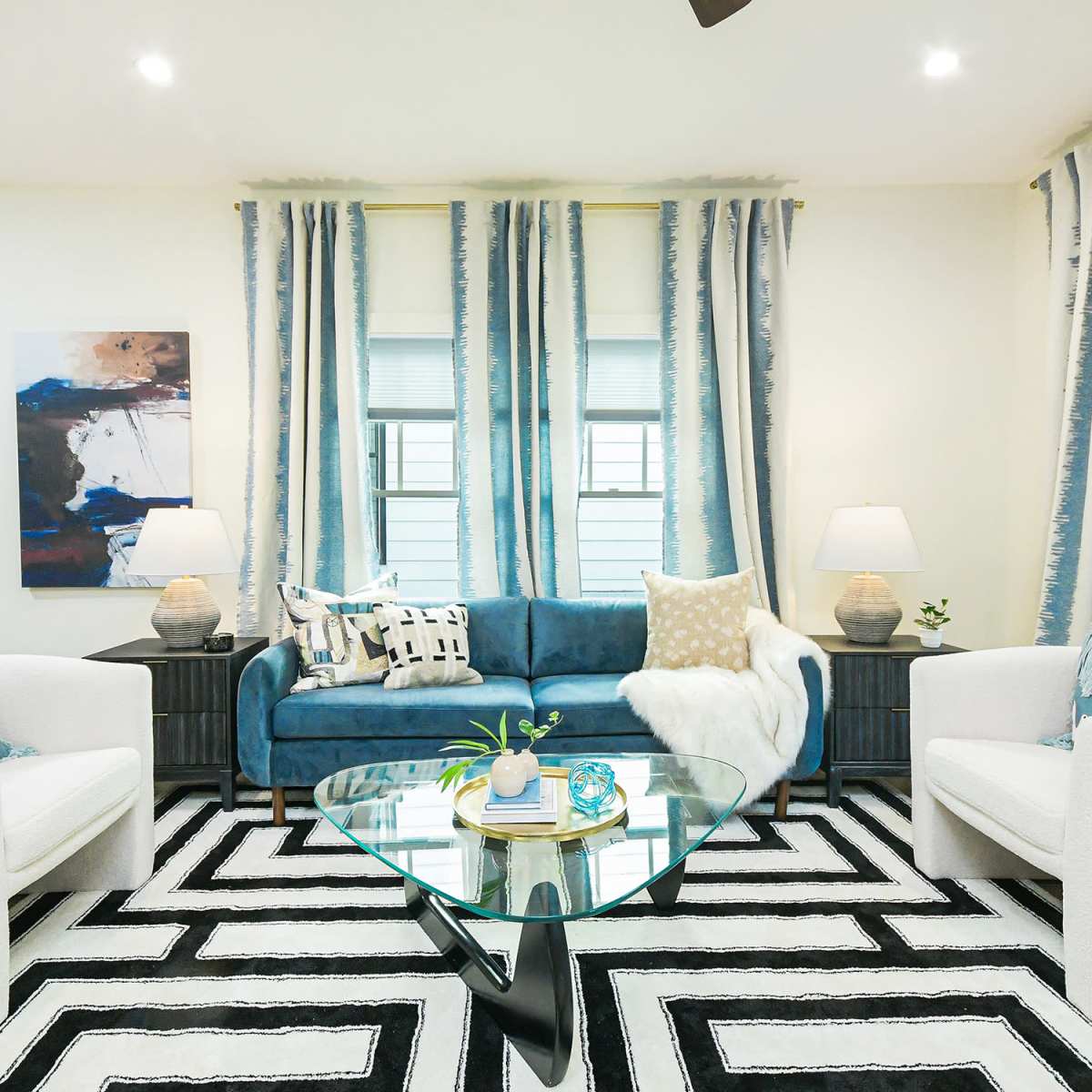A modern living room features a blue velvet sofa, two white armchairs, a glass coffee table, and striped curtains, all arranged on a black-and-white rug.