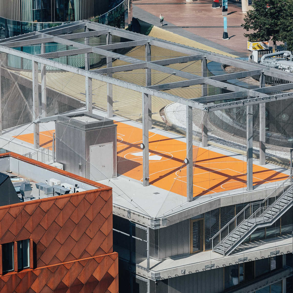 A rooftop basketball court with an orange playing surface and a protective netting, surrounded by modern buildings.