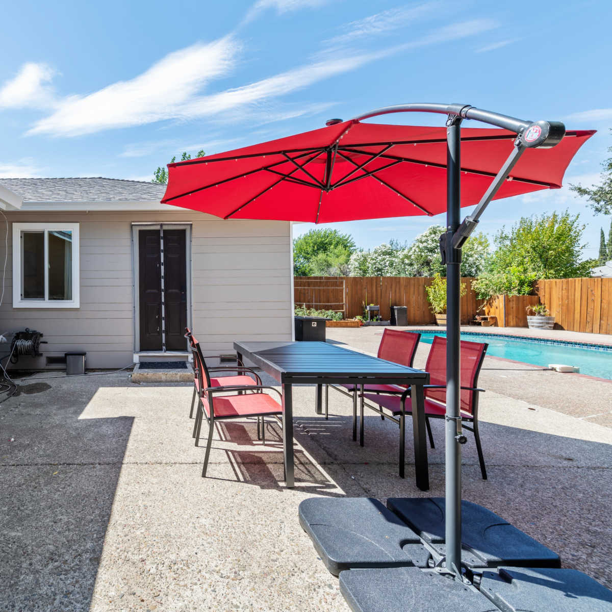 A backyard patio area with a table and chairs under a red umbrella beside a house and a pool.