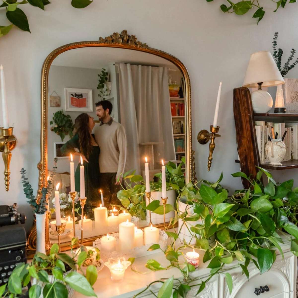 A couple stands in front of a mirror adorned with candles and plants in a cozy room.