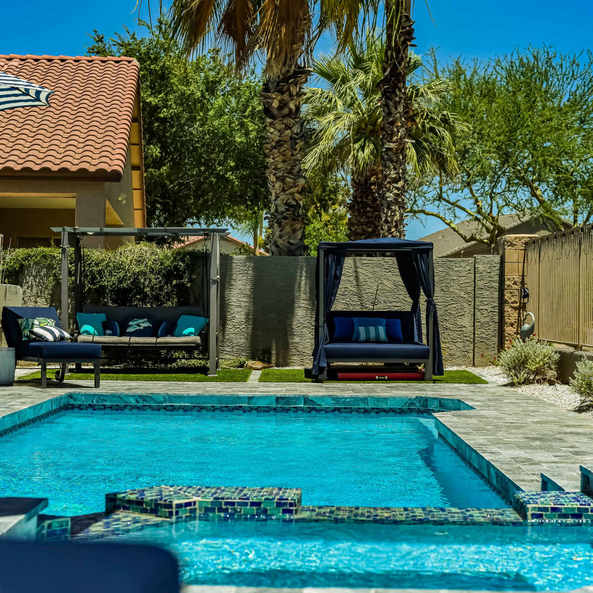 A residential backyard featuring a swimming pool, lounge chairs, a swinging daybed, and palm trees under a clear blue sky.