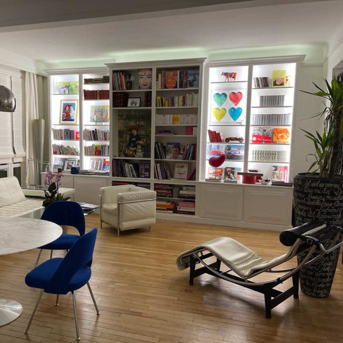 A modern living space features a circular marble dining table surrounded by blue chairs, with white sofas and a large bookshelf displaying colorful artwork and decor.