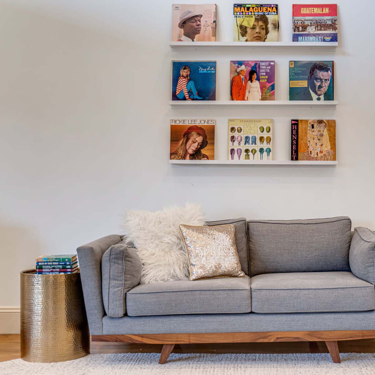 A gray sofa with decorative pillows sits in front of a wall-mounted shelf displaying various vinyl records, alongside two metallic side tables.