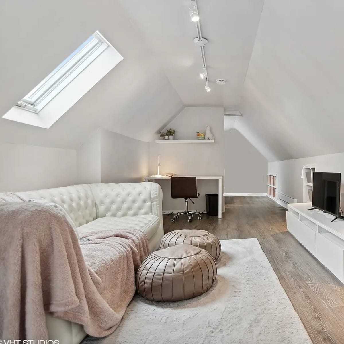 A modern attic space with a white sofa, a desk with a chair, a TV stand, and two large cushioned poufs.