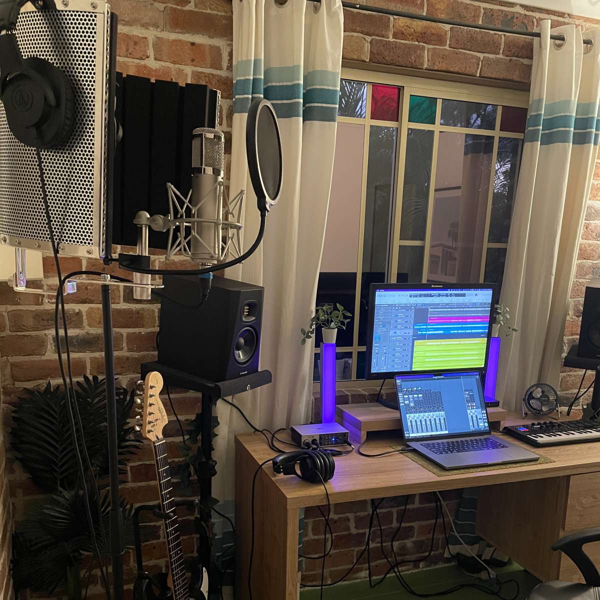 A home music studio features a microphone setup, computer with audio software, and various instruments arranged around a wooden desk.