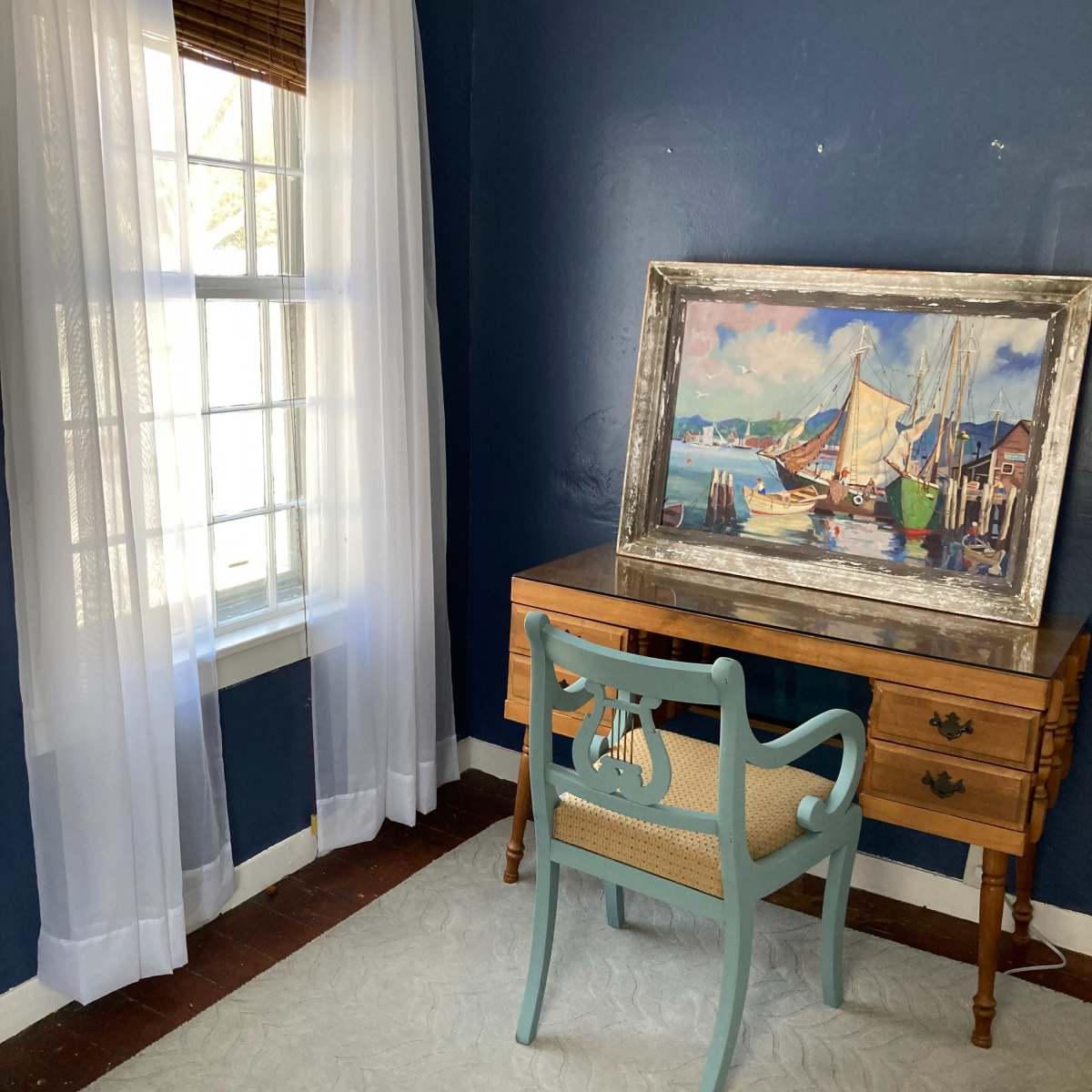 A small room with blue walls, a wooden desk, a light blue chair, and a framed painting of boats on the wall beside a window with sheer white curtains.