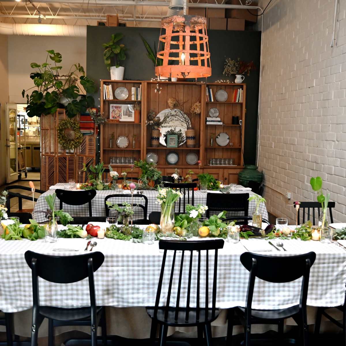 A long dining table is set with a checked tablecloth and arranged with fruits, greenery, and candles in a room featuring plants, shelves, and decorative items.