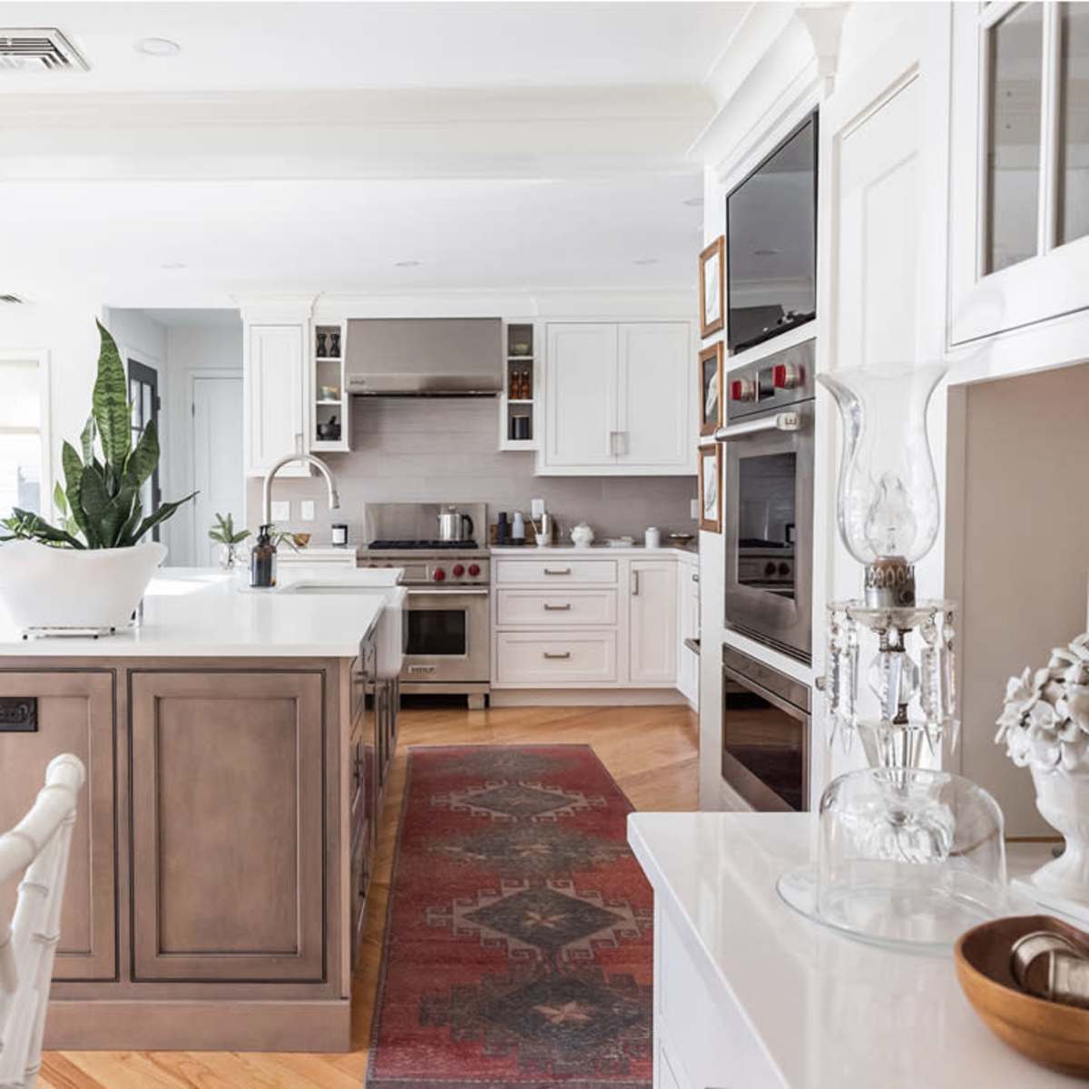 A modern kitchen with an island, featuring white cabinets, a stove, and a decorative plant, beside a dining area with a round table and upholstered chairs.