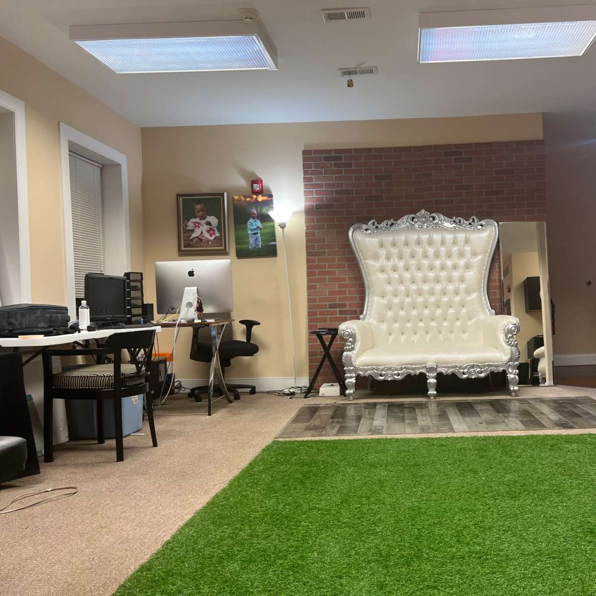 A spacious room with two ornate white chairs, a green area rug, a desk with a computer, and a cozy black couch in a well-lit environment.