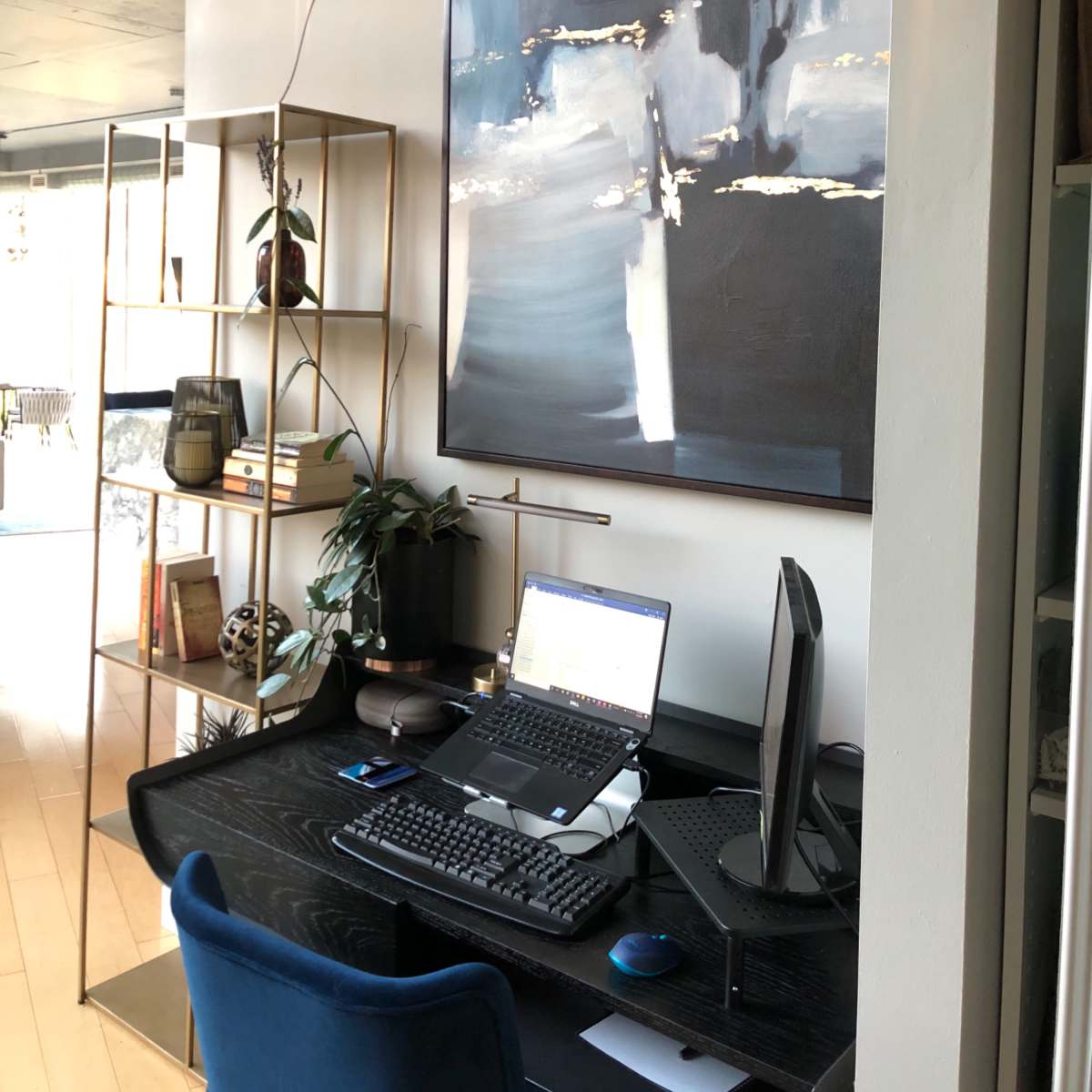 A modern office space features a wooden desk with a laptop, monitor, and keyboard, accompanied by a bookshelf and a large abstract painting on the wall.