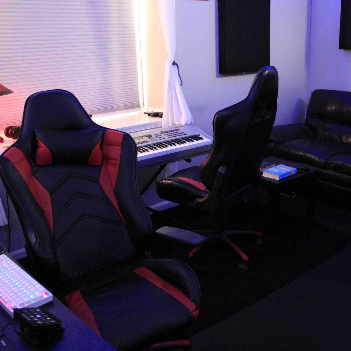 A music studio setup with two gaming chairs, a keyboard on a desk, and a black leather couch against the wall.