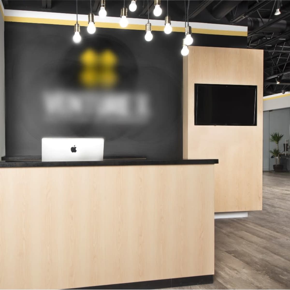 A sleek reception desk made of light wood with a black countertop, featuring a computer on it, set against a dark wall with decorative lighting and a television.