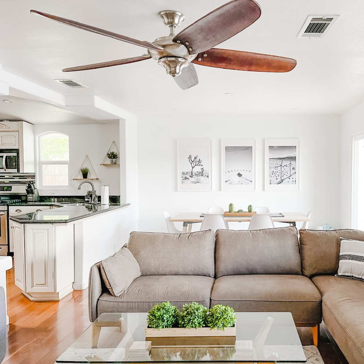 A bright living room features a sectional sofa, a glass coffee table with greenery, and a ceiling fan overhead, with a dining area visible in the background.
