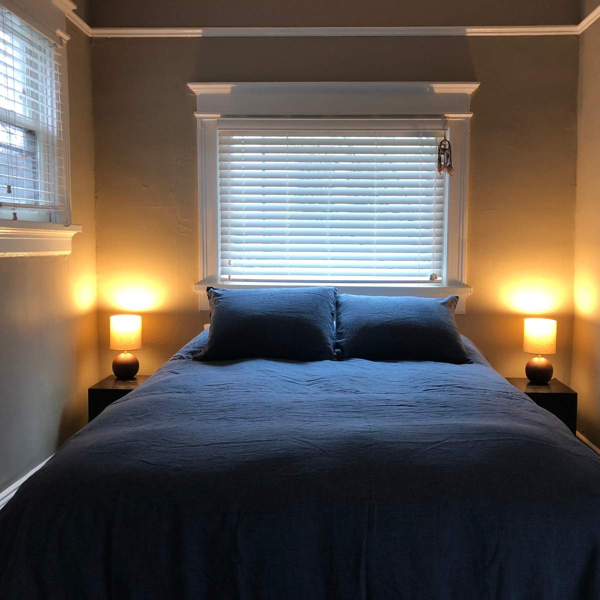 A neatly made bed with blue bedding is positioned between two bedside tables with illuminated lamps beneath a window with closed blinds.