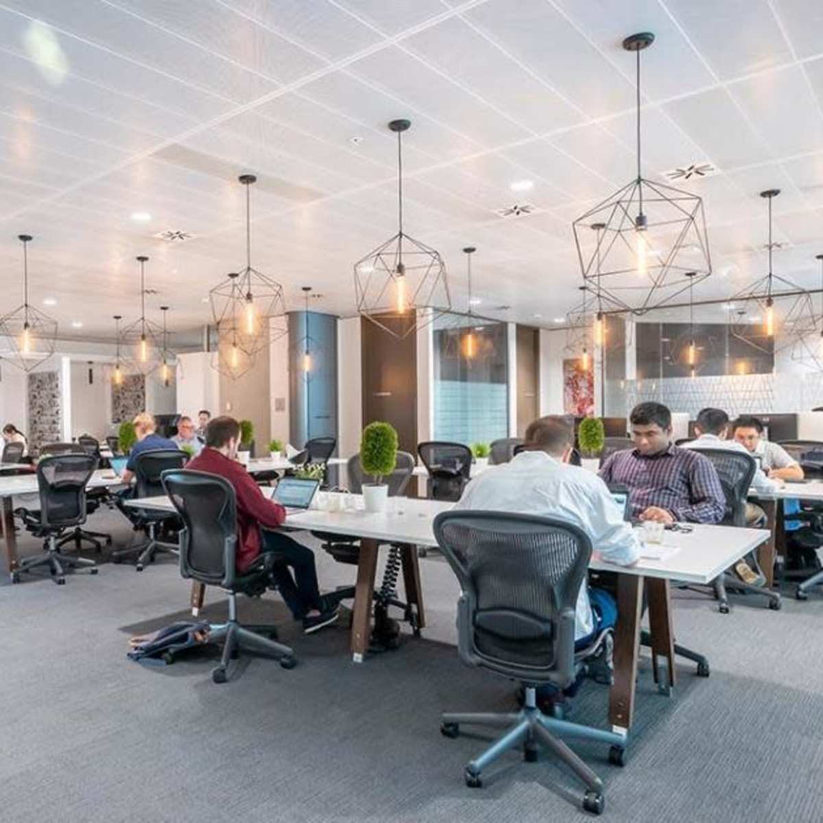 A modern office space with several workers seated at desks, utilizing computers under geometric pendant lighting.