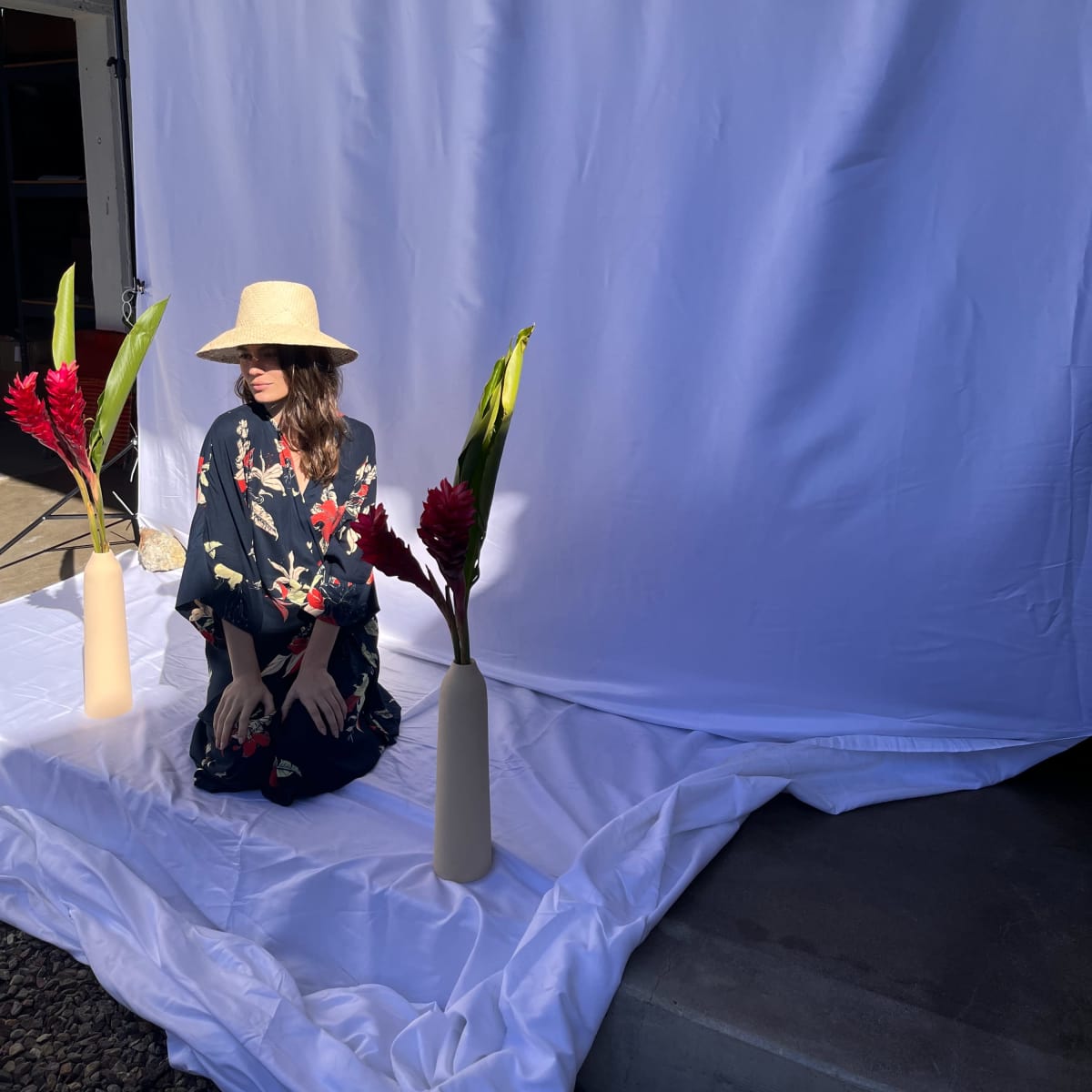 A woman in a floral outfit sits in front of a white backdrop with two vases containing flowers on either side.