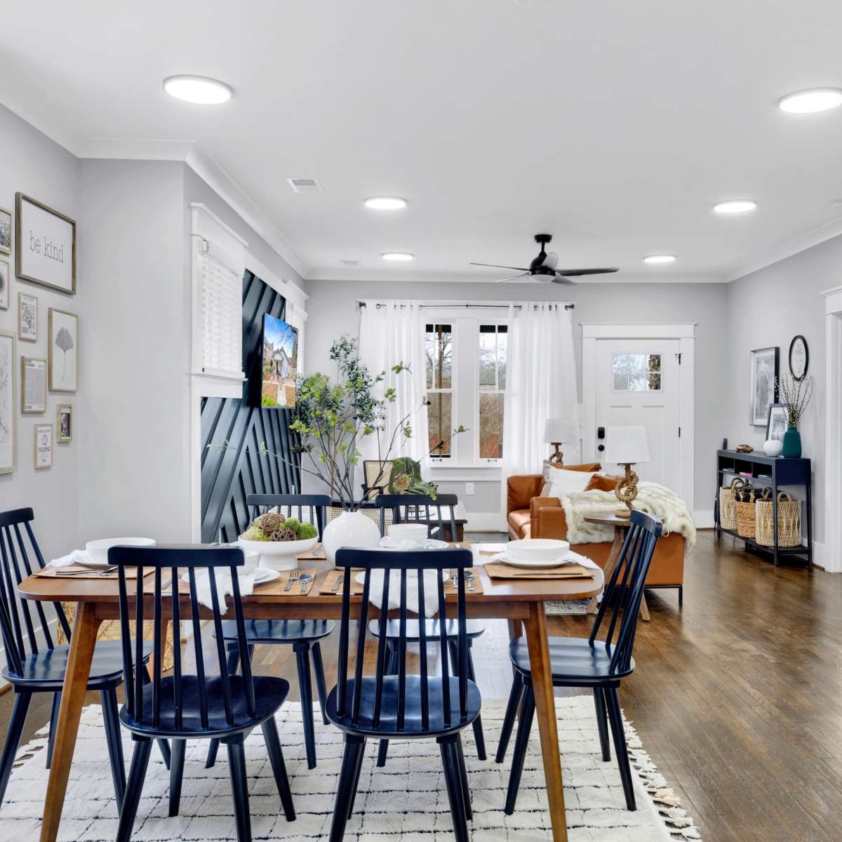 A modern dining area features a wooden table set for meals, surrounded by dark blue chairs, with a living room visible in the background.